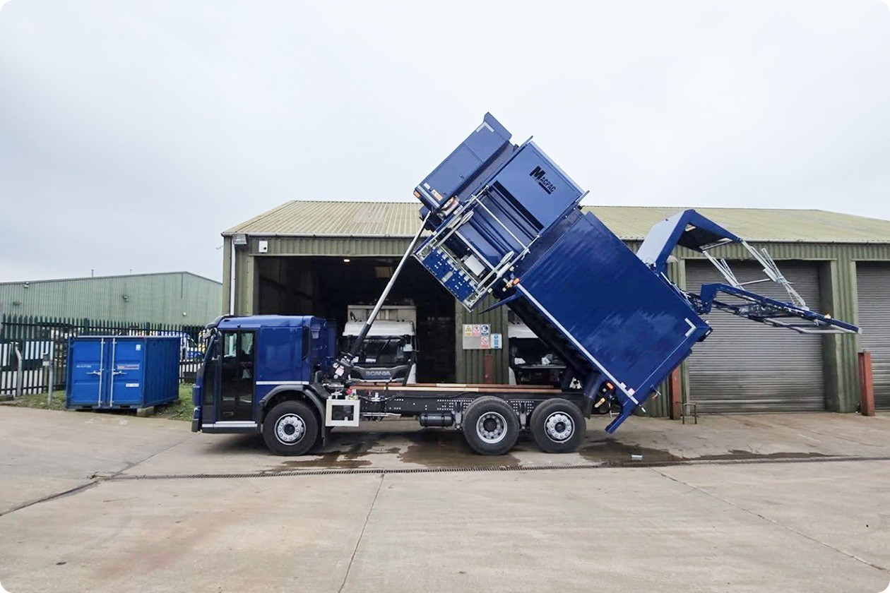A blue dump truck is positioned next to a building, with its bed raised for unloading.