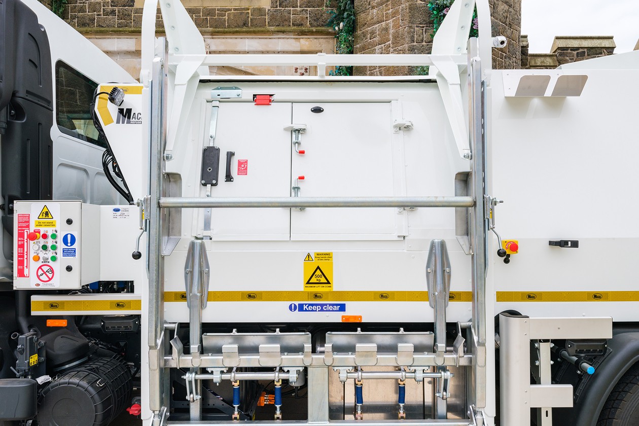 A rear view of a garbage truck with its rear loading compartment visible, showcasing warning labels and operational controls.