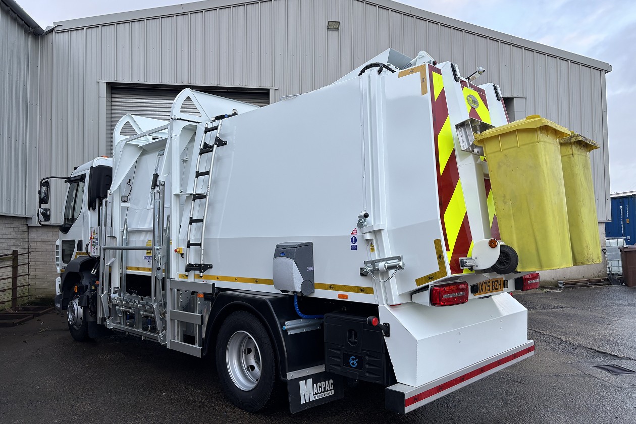 A white garbage truck parked outside a warehouse, with a yellow waste bin attached to the back.