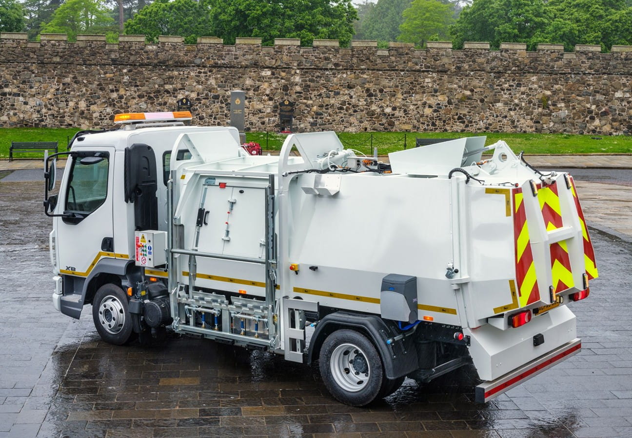 A white garbage truck with reflective safety stripes, parked on a wet stone pavement in front of a stone wall.